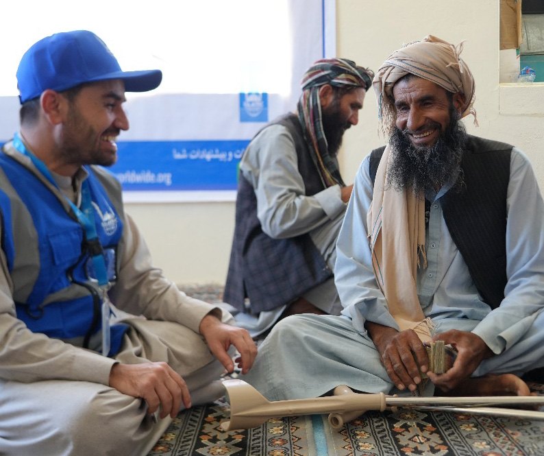 An Islamic Relief staff member in a blue cap and vest sits on a patterned floor mat talking and laughing with an older man in a turban, who is holding a bundle of banknotes.