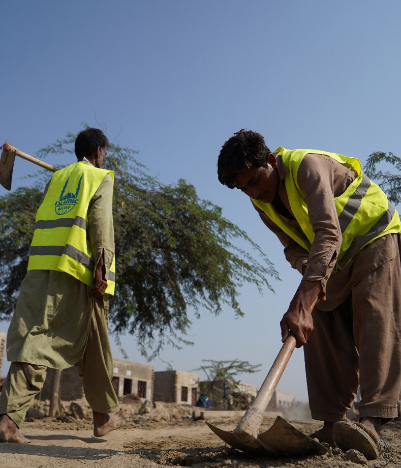 Two men in high-visibility Islamic Relief vests dig with shovels to clear ground beside a damaged road in Pakistan.