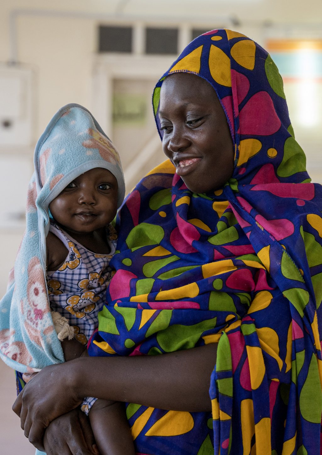 Baby Sarah, wrapped in a pale blue blanket, held by a woman in a brightly patterned headscarf at a healthcare centre in Gedaref, Sudan.