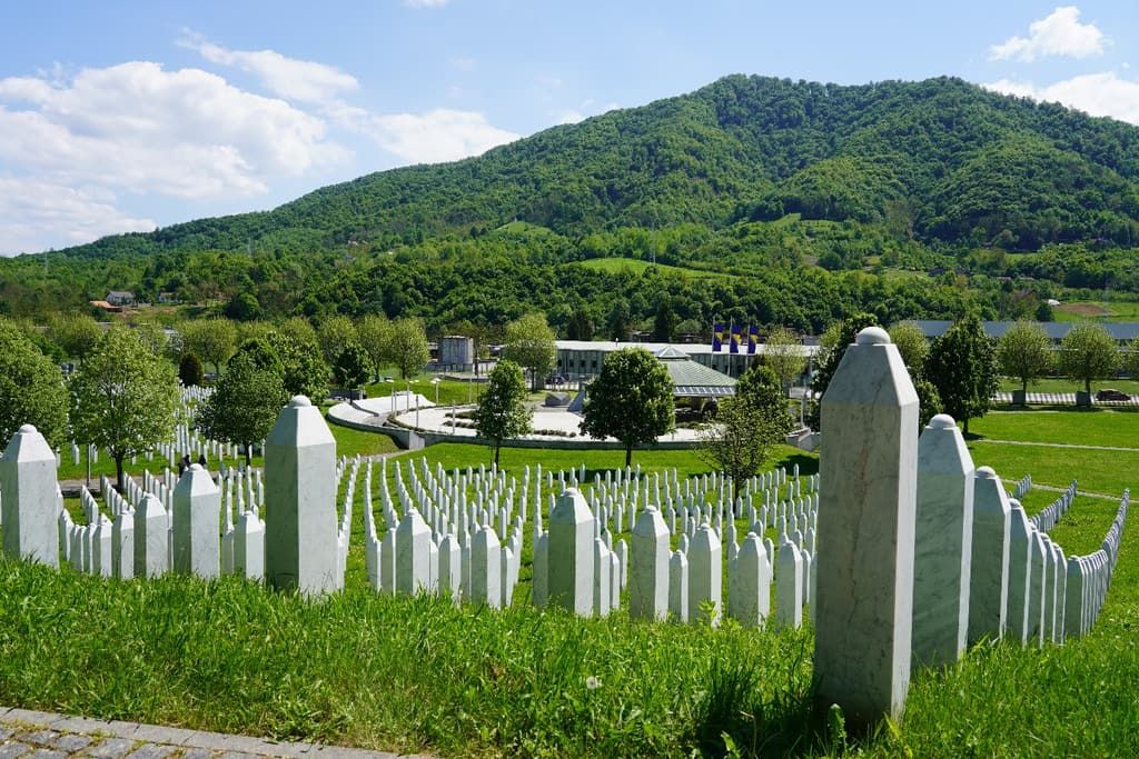 Rows of white marble grave markers at the Srebrenica–Potočari memorial cemetery, with green hills behind.
