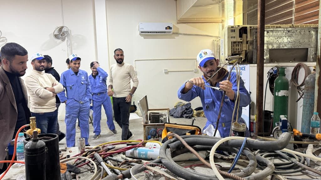 Trainees in blue overalls watch a welding demonstration in a vocational training workshop in Iraq.