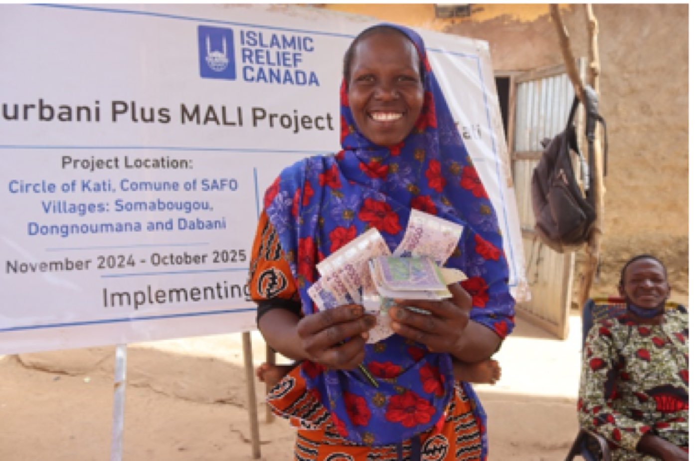 A woman smiles while holding a cash grant in front of a Qurbani Plus Mali Project banner.