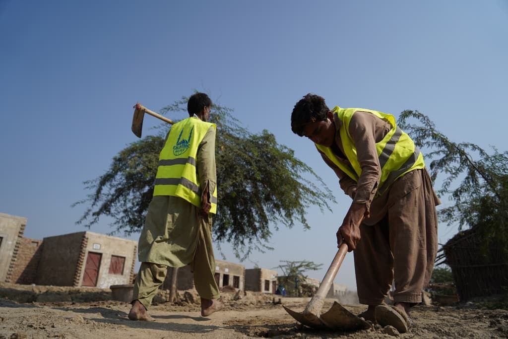 Two men in high-visibility vests clear ground with hand tools during a cash-for-work scheme in flood-affected Pakistan.