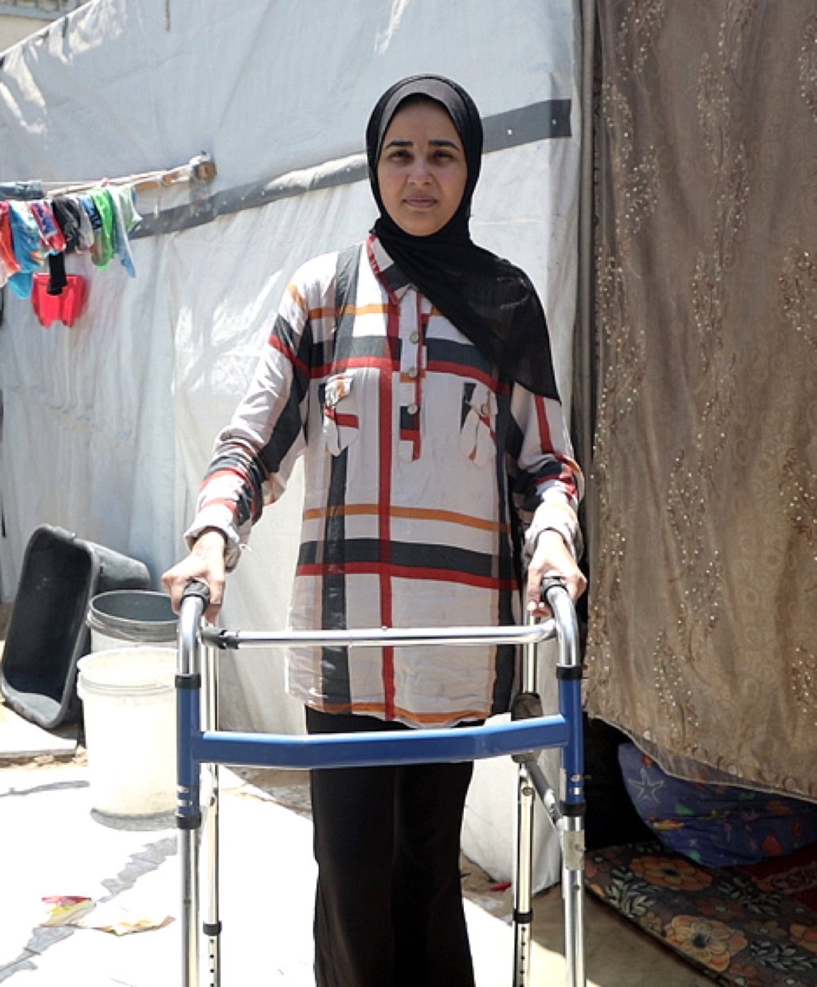 Rama, a teenage girl in a headscarf, stands supported by a walking frame outside a tent in a displacement camp.