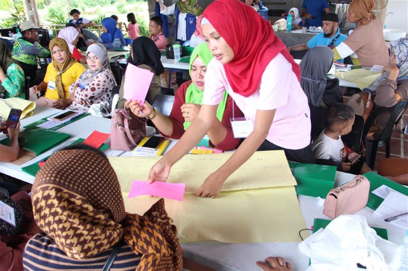 Women take part in a community workshop, sorting coloured cards on tables under an open shelter.