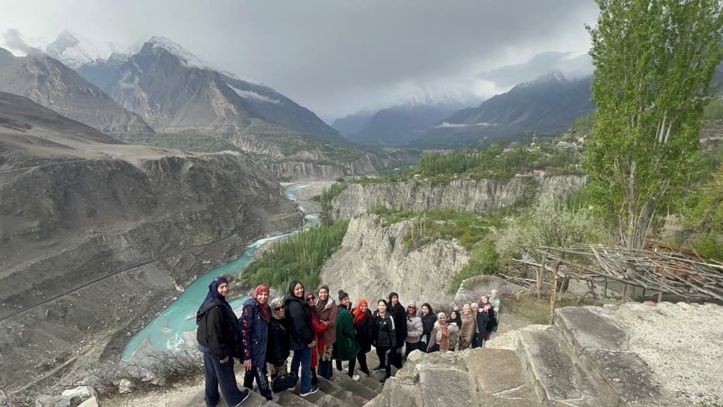 A group of trekkers stands on a rocky ridge above a turquoise river in the mountains of northern Pakistan.