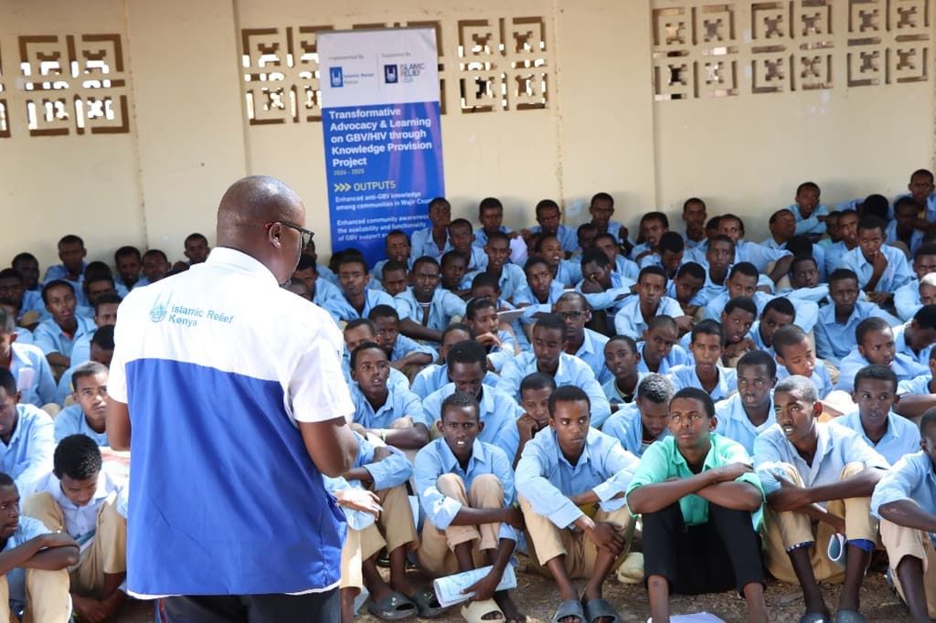 An Islamic Relief staff member in a branded gilet addresses a large group of seated schoolchildren in a school courtyard in Kenya.